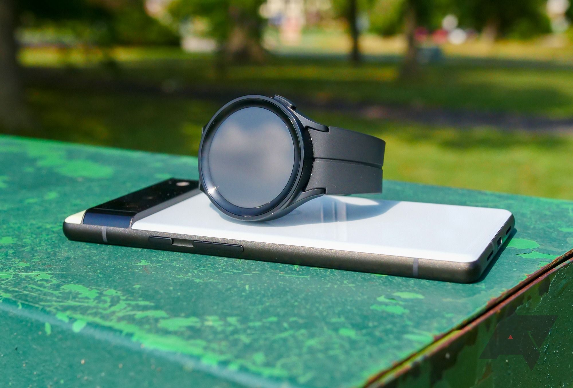 A smartwatch sitting on top of a smartphone on a table in a park.