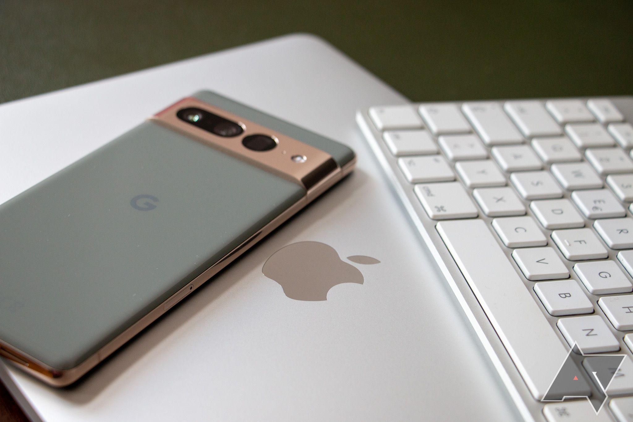 A Google Pixel 7 Pro laying on top of a MacBook Pro, with the Apple emblem visible. A wireless Mac keyboard is placed at the valid.