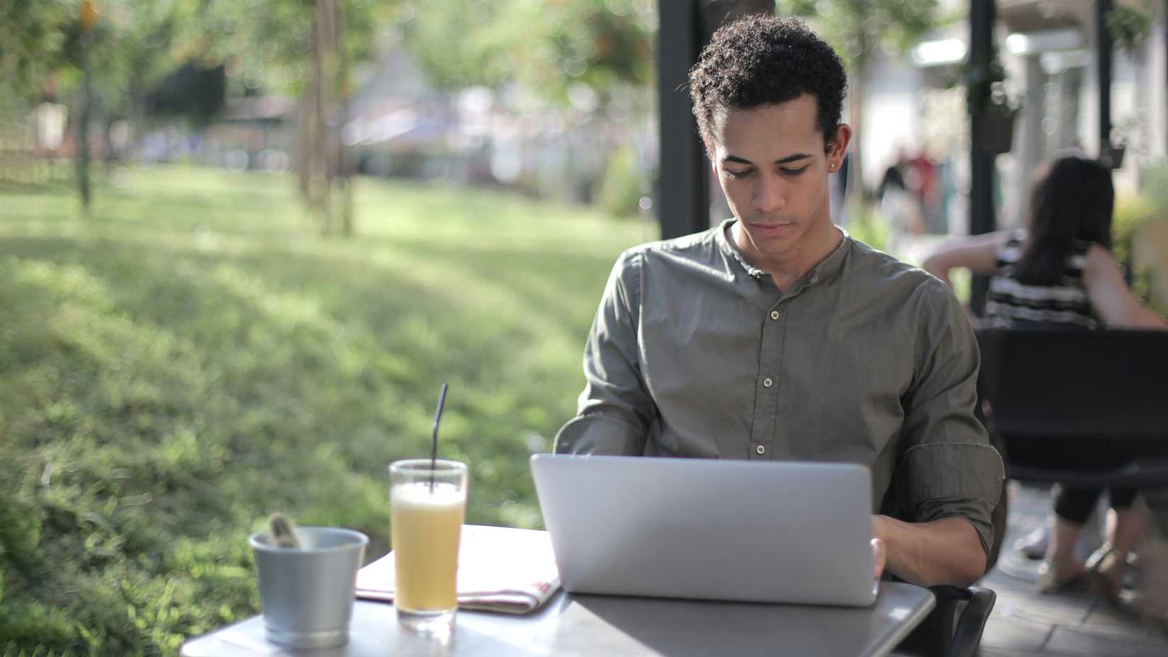 شخص يجلس في الخارج مع مشروب وينظر إلى جهاز كمبيوتر محمول فضي.