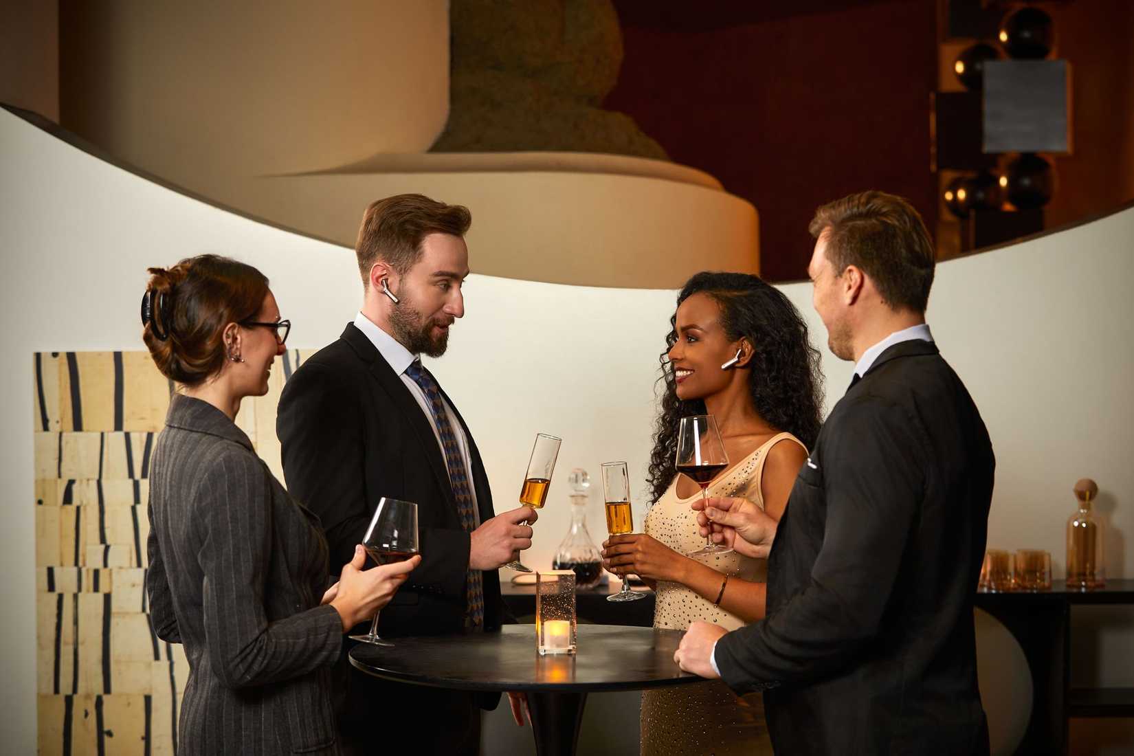 Four people drinking cocktails and talking while two wear Timekettle AI Interpreter earbuds.