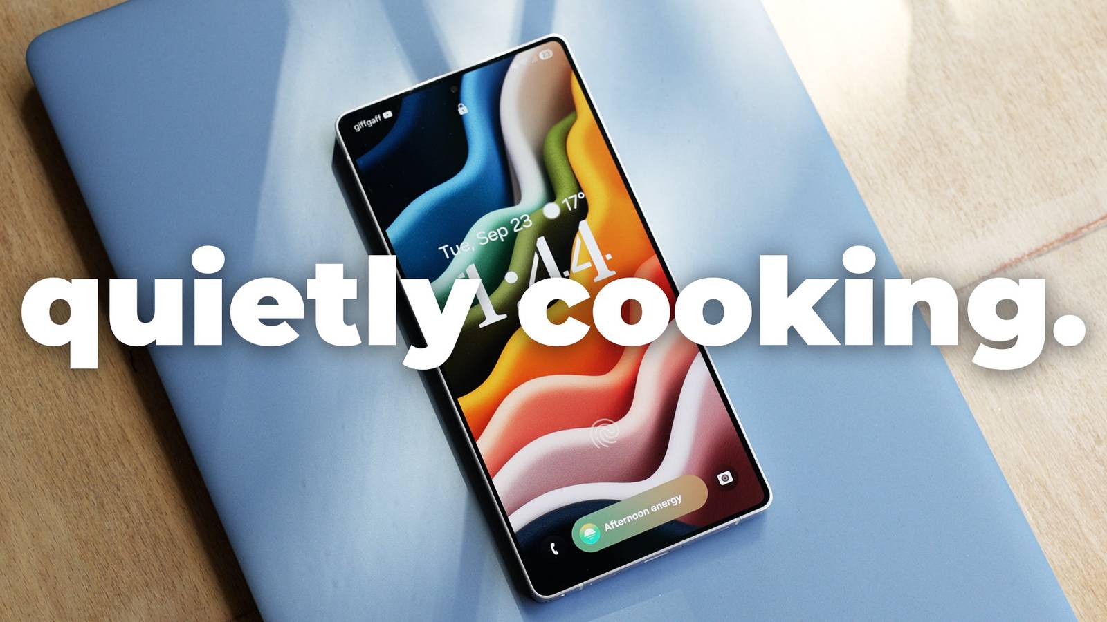A smartphone with a colorful wavy wallpaper sits on a light blue laptop, with the bold text “quietly cooking.” overlaid on the image