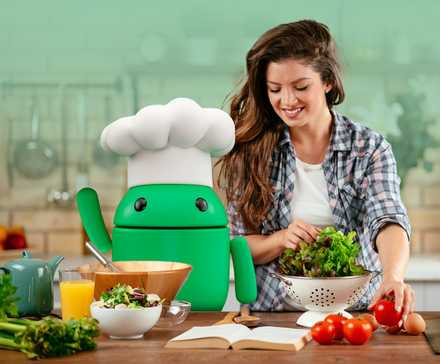 Mulher sorridente preparando uma salada em uma cozinha ao lado do mascote Android usando chapéu de chef, cercada por vegetais frescos e itens de cozinha