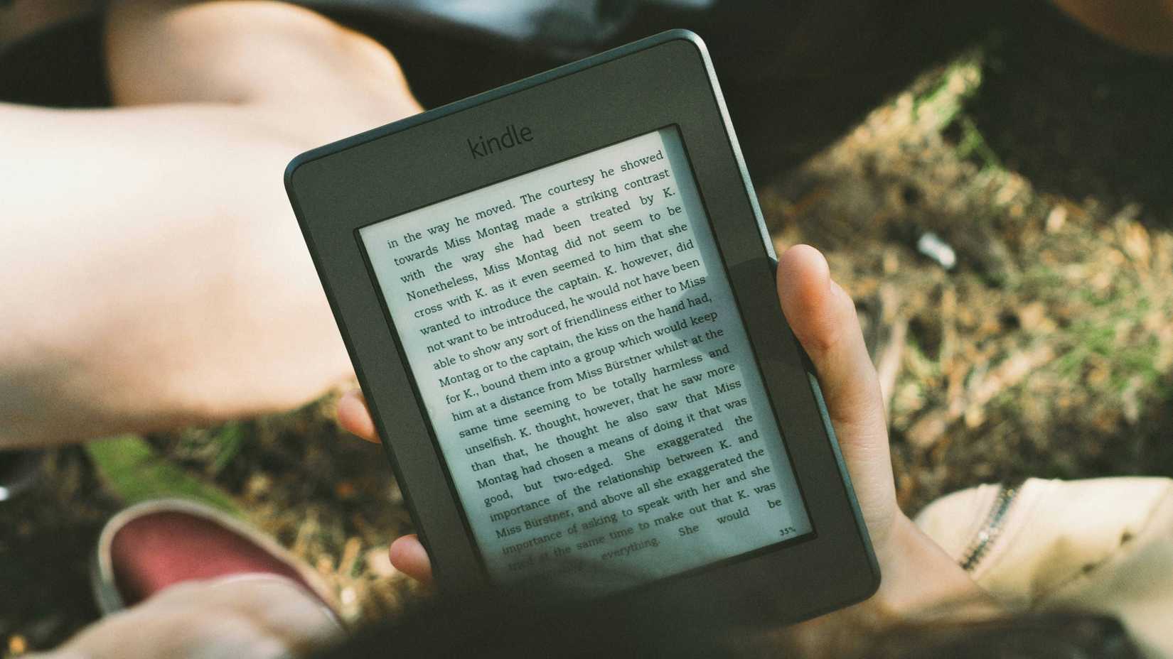 A kindle e-reader displaying text, being held by a person sitting in grass