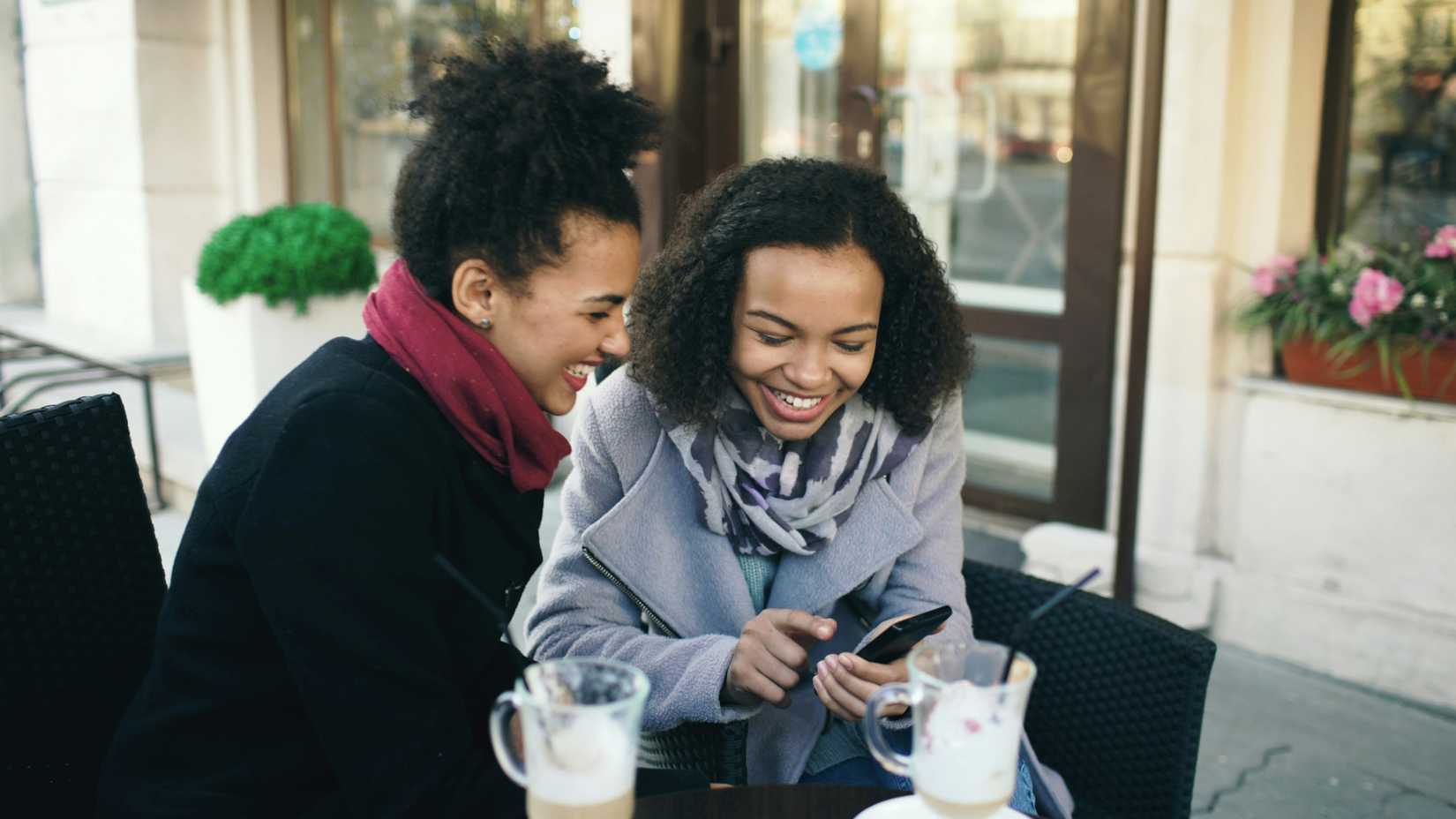 A photo of two women laughing at a smartphone over coffee