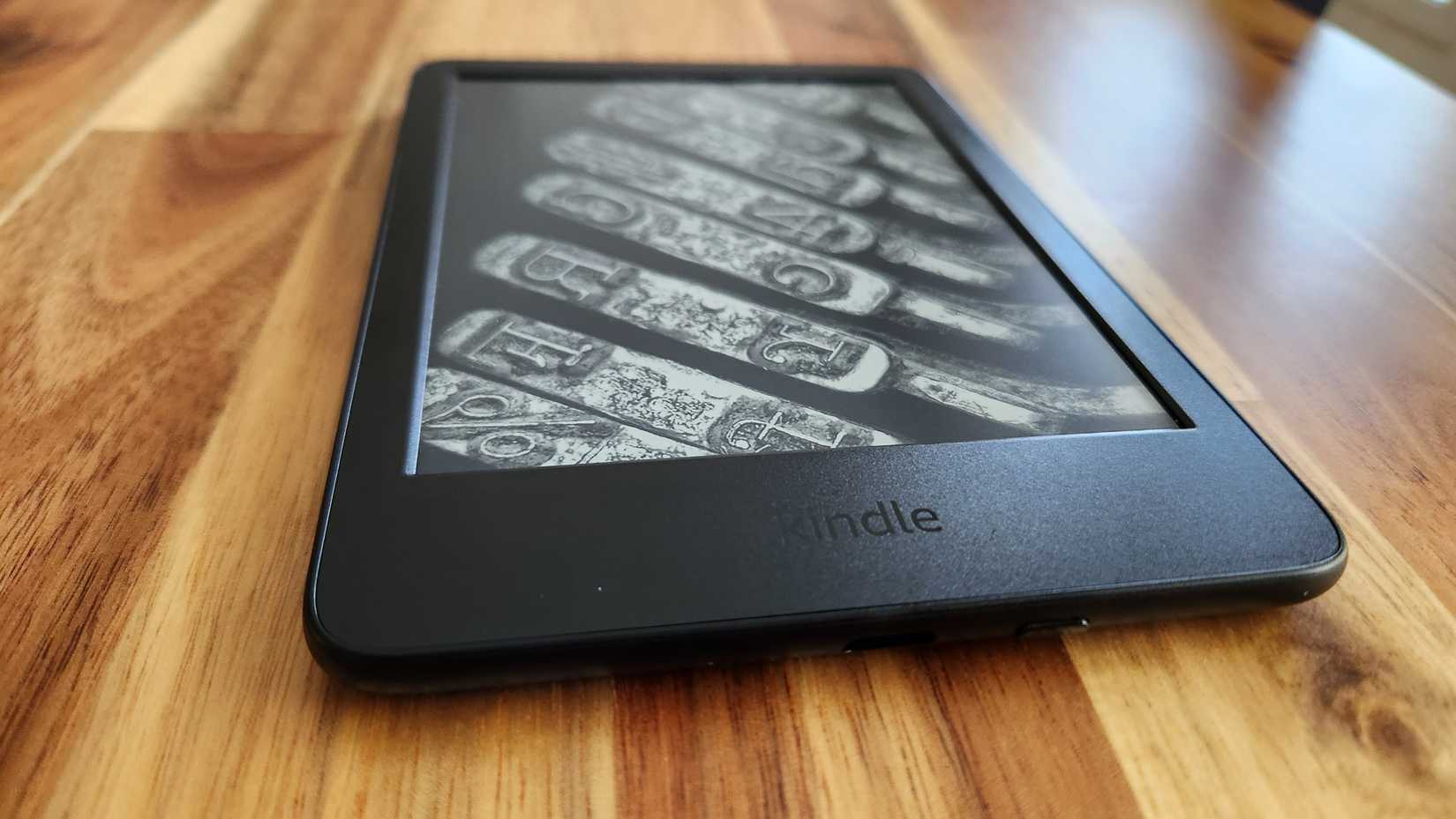 An Amazon Kindle E-Reader sits on a wooden table.  It's screen is a lock screen picture of old fashioned typewriter keys.