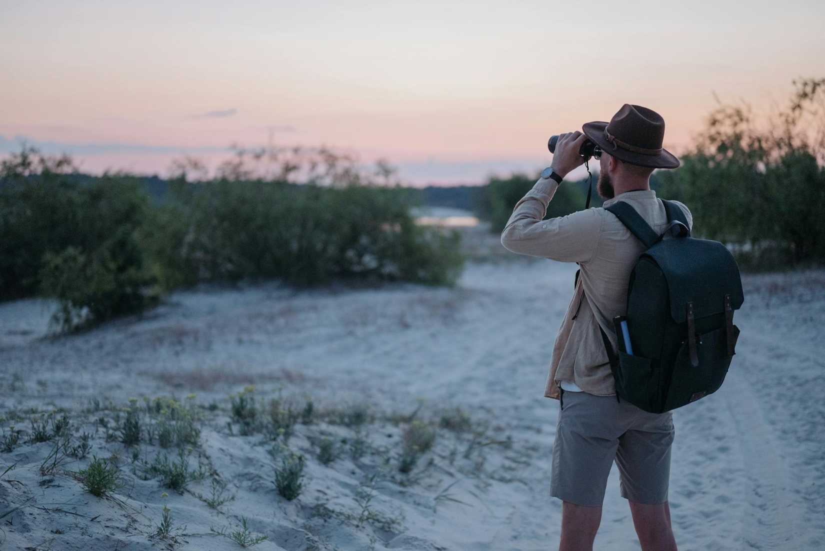 Homem com mochila observando a natureza através de binóculos durante um pôr do sol cênico.