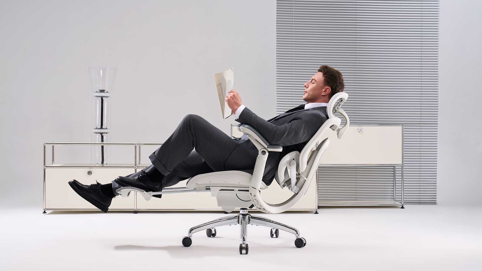 Um homem de terno preto reclina-se lendo jornais em uma cadeira ergonômica branca