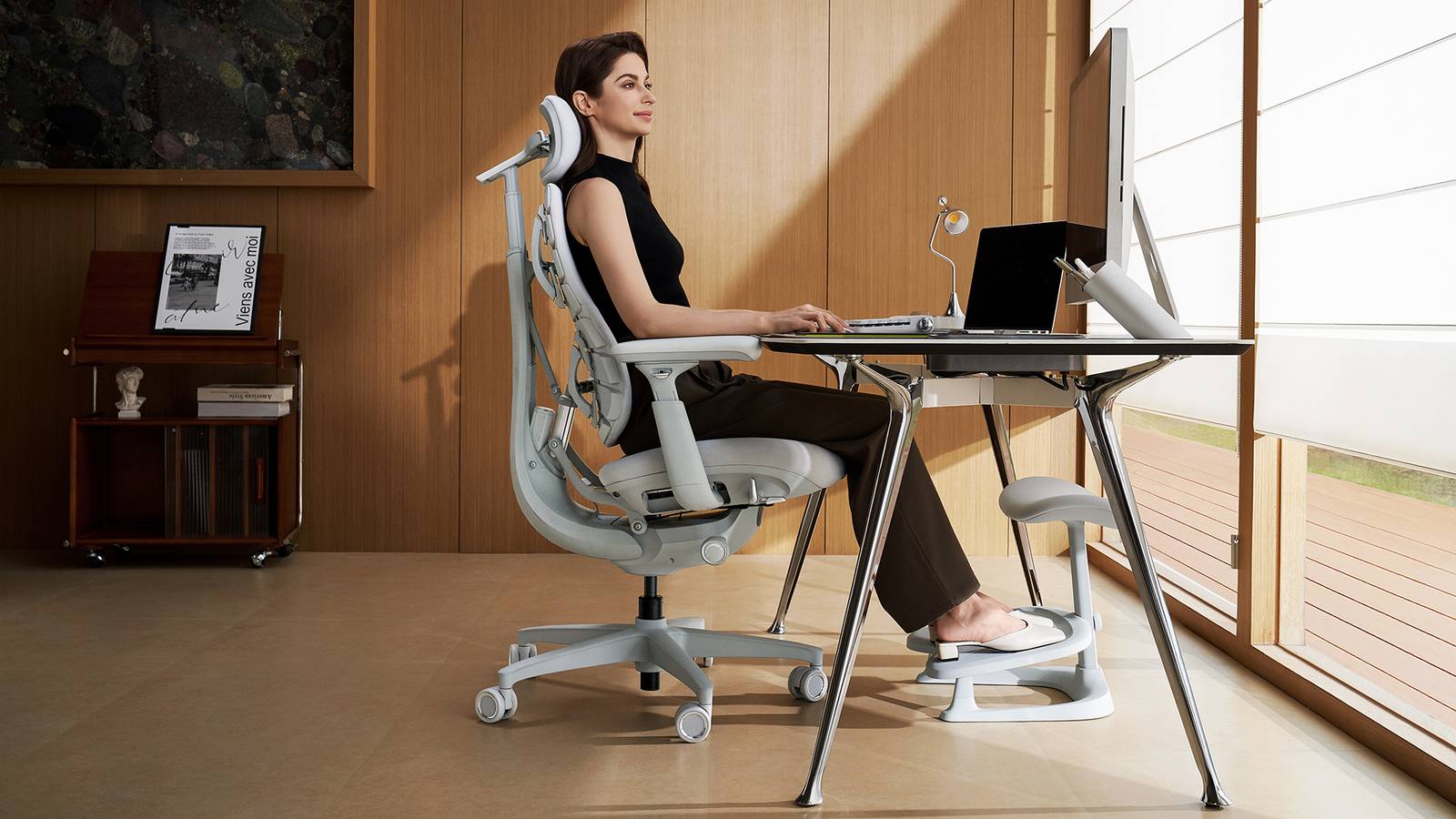 A woman sits at a computer desk in a grey ergonomic desk chair.