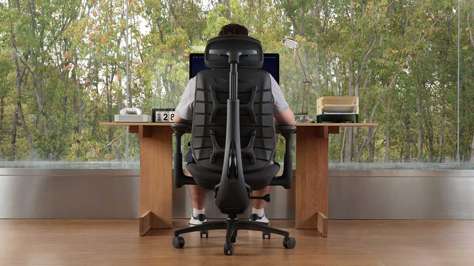 A man sits in a black ergonomic chair at a desk overlooking a window in front of the forest.
