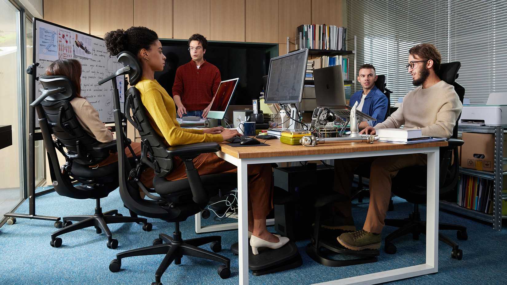 Four people sit in matching ergonomic chairs in an office while one man stands and talks.