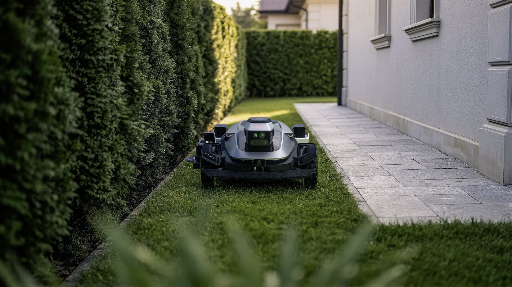 Front view of the Sunseeker X8 Plus robotic lawn mower between hedges and a home.