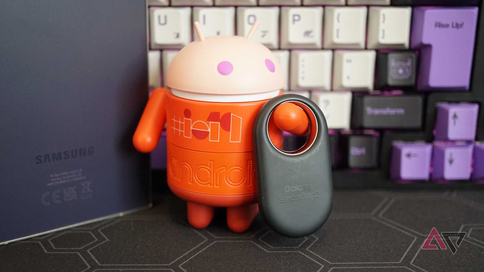 Uma foto de um Android segurando o Galaxy SmartTag 2 com teclado e S24 Ultra em segundo plano