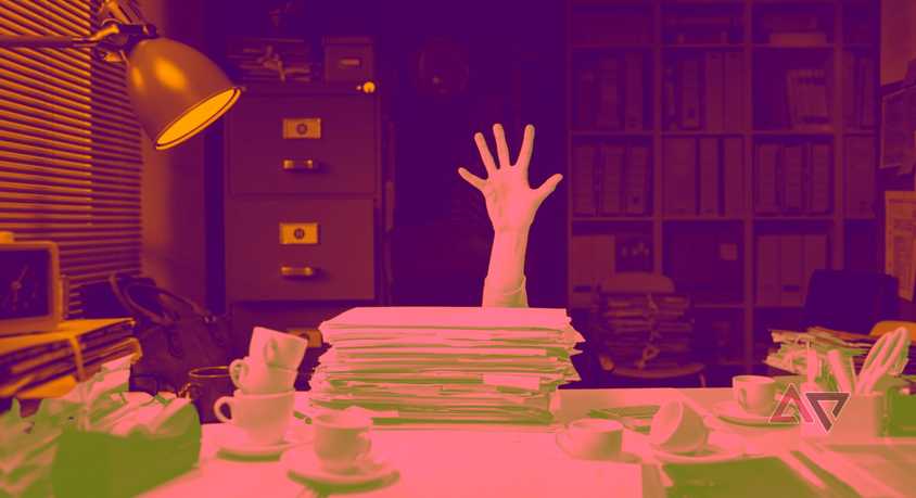 A hand grasping at the air in desperation over a stack of papers as its owner sinks behind a cluttered desk