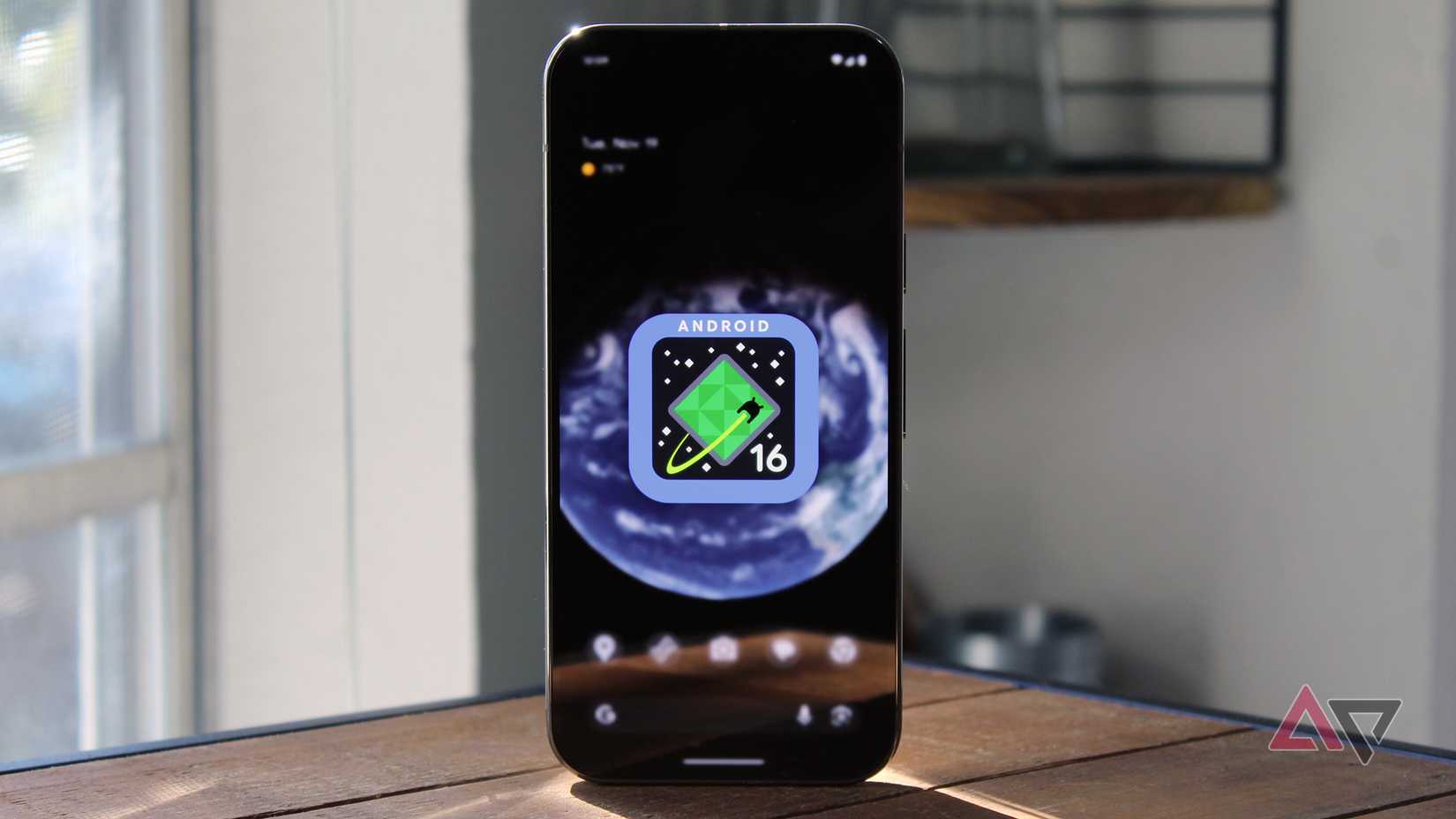 A tela inicial do Pixel 9 Pro apresenta uma imagem do logotipo do Android 16 sobre uma mesa de madeira.