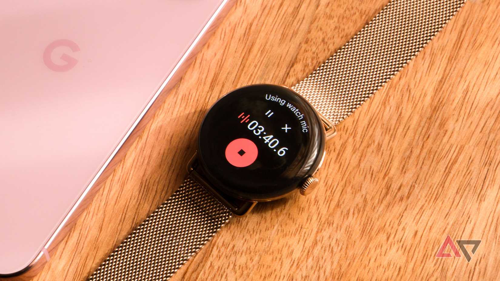 O aplicativo Google Pixel Watch 3 com gravador na tela fica em uma mesa de madeira ao lado de um telefone Google Pixel rosa.