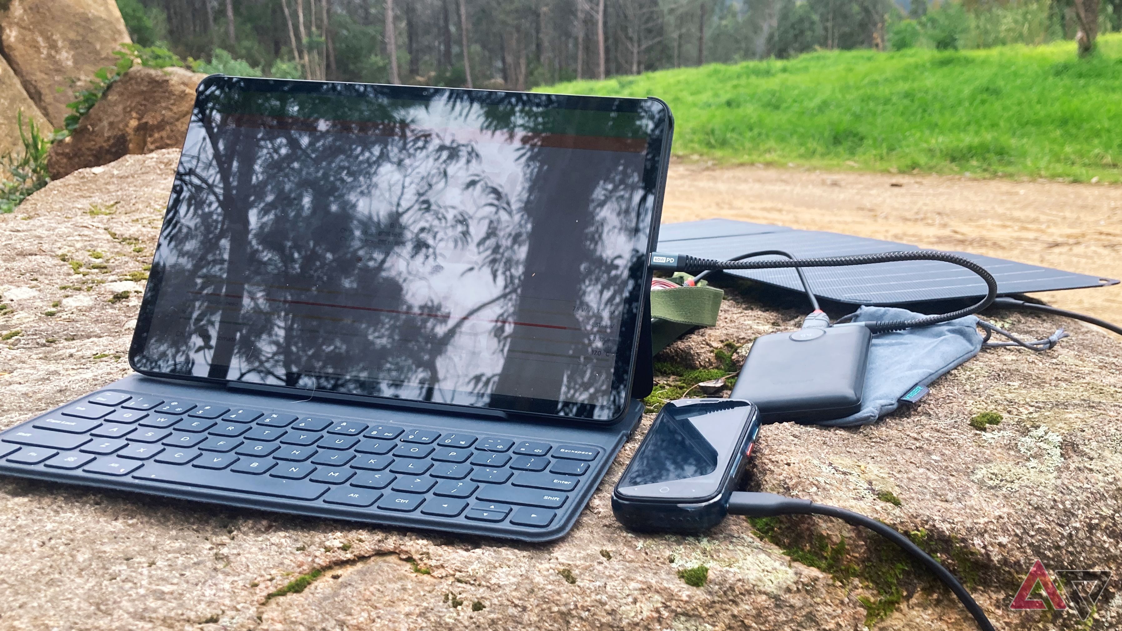 Custodia per tablet e tastiera, smartphone, power bank, pannello solare e un assortimento di cavi su una roccia all'aperto in un ambiente naturale.
