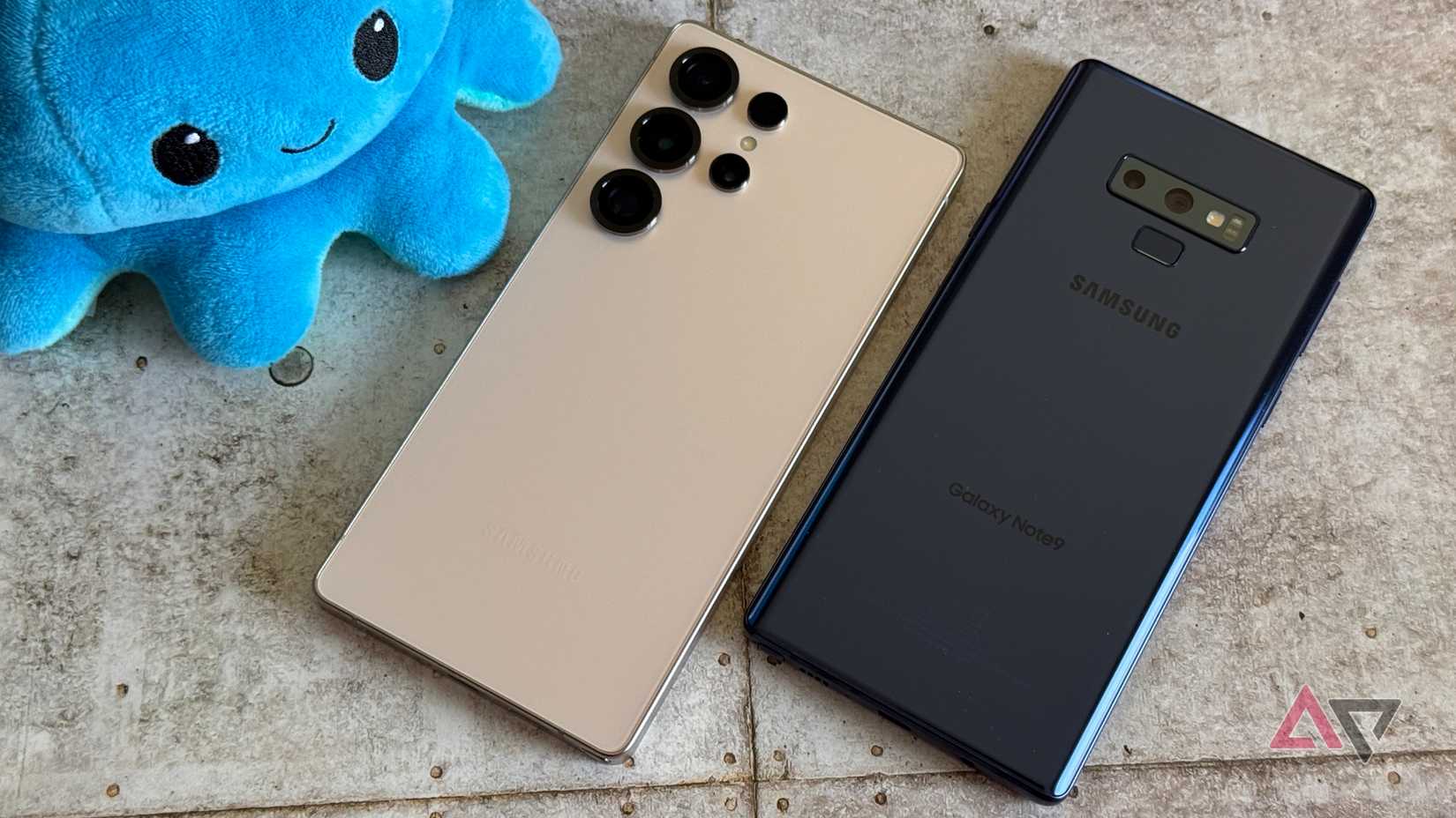 Un Samsung Galaxy Note 9 e Galaxy S25 Ultra uno accanto all'altro su un pavimento piastrellato con un peluche blu