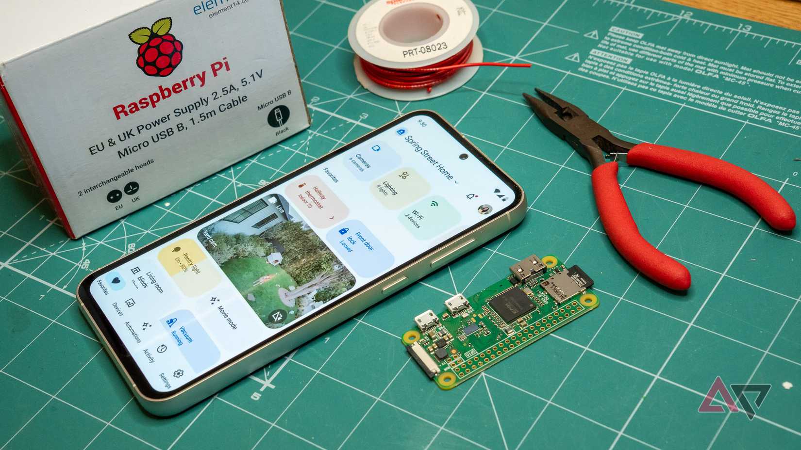 Um telefone exibindo o aplicativo do Google Home em um tapete verde ao lado de alicate, arame, um Raspberry Pi e uma caixa branca