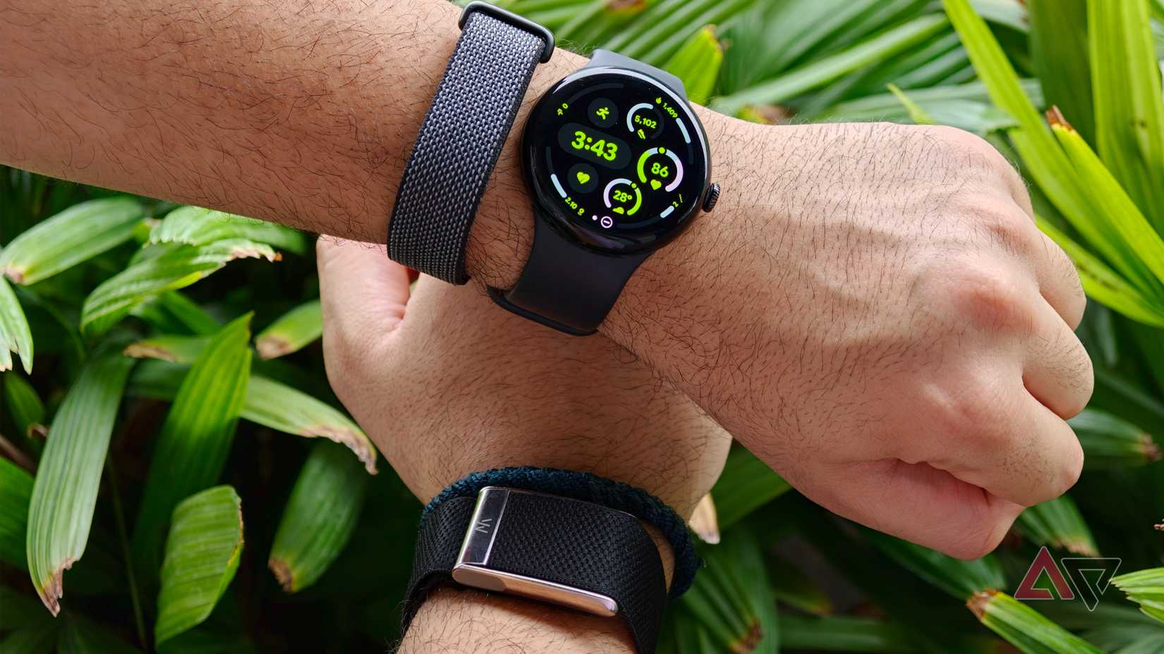 Two wrists positioned together, one wearing a round Google Pixel Watch with a grey stretch band, and the other wearing a screenless Whoop fitness tracker with a black fabric band