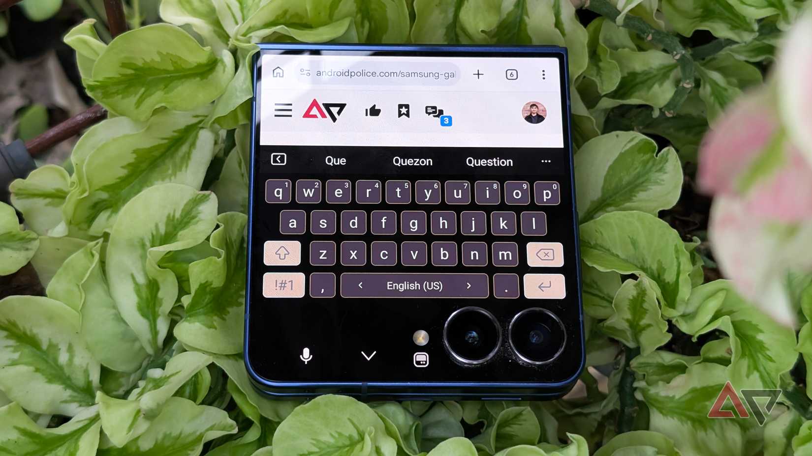 Samsung Keyboard app on Galaxy Z Flip 7 smartphone on top of a plant and with Android Police website shown