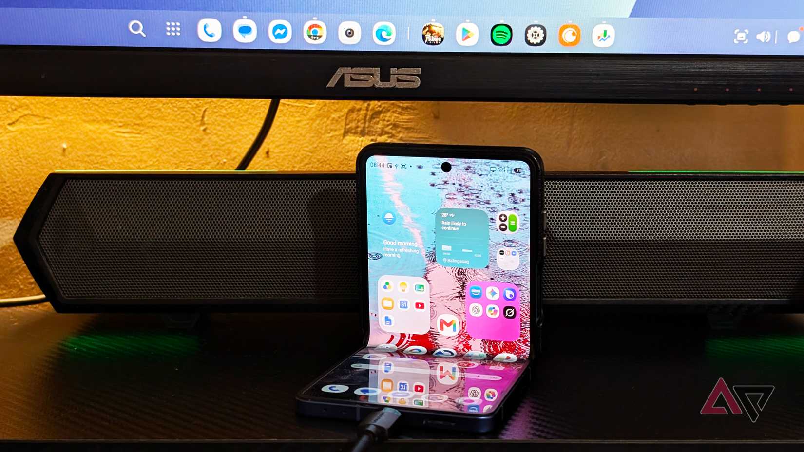A Samsung Galaxy Z Flip smartphone halfway folded on a desk with a monitor on the back